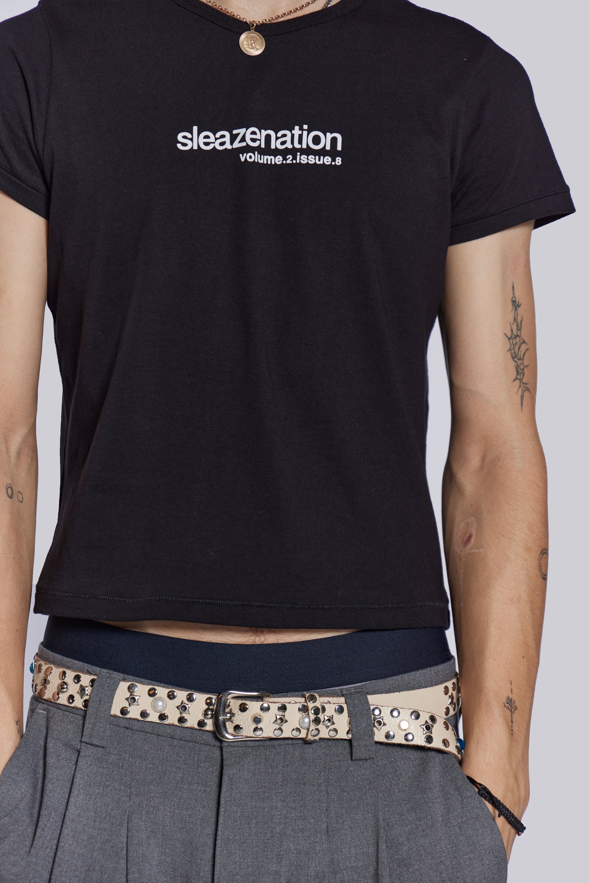 Male wearing black slogan t-shirt, styled with grey pleated trousers and studded belt.