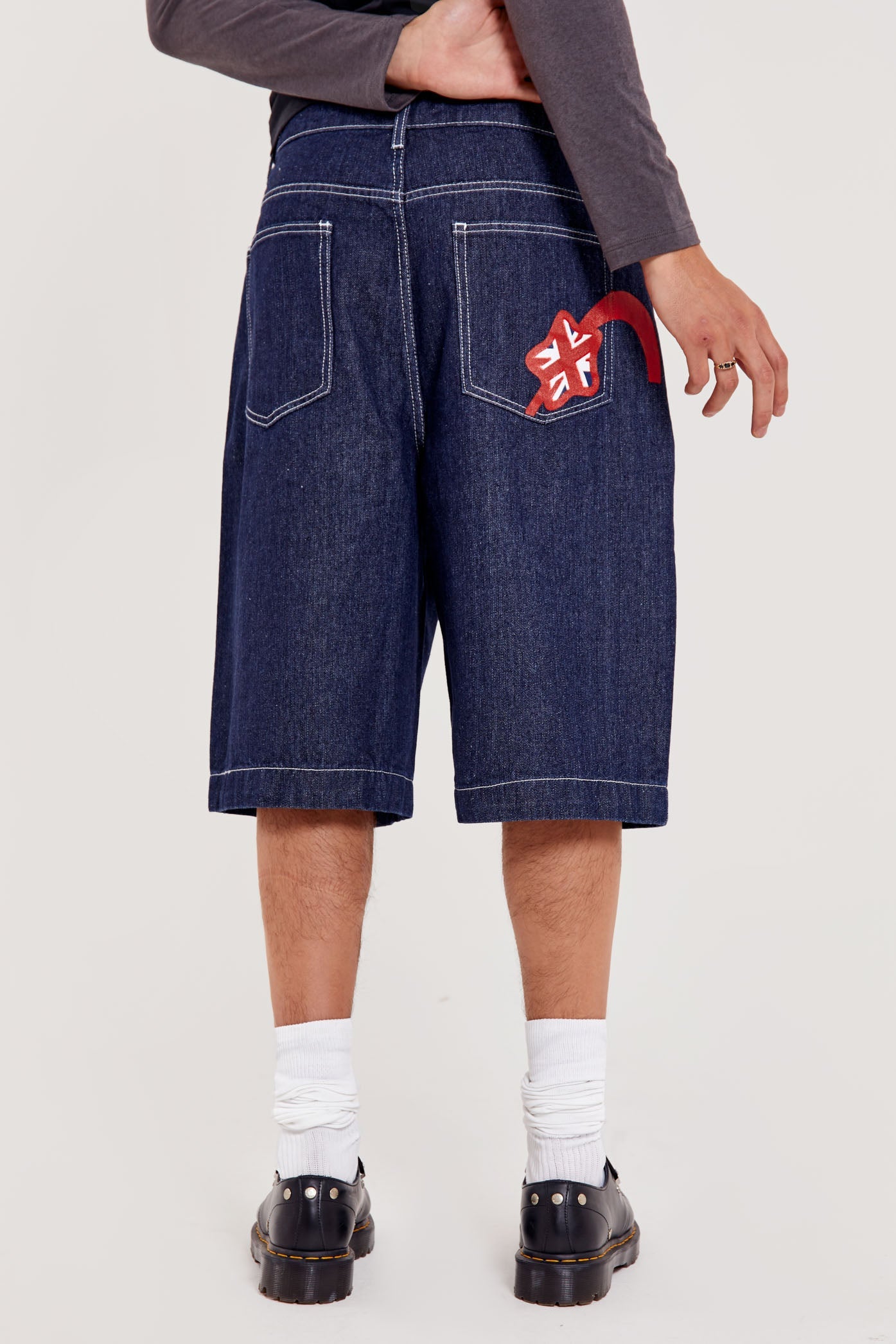 Male wearing indigo blue denim jorts in a jumbo fit with JL embroidered logo and Union Jack start branding on back pocket. 