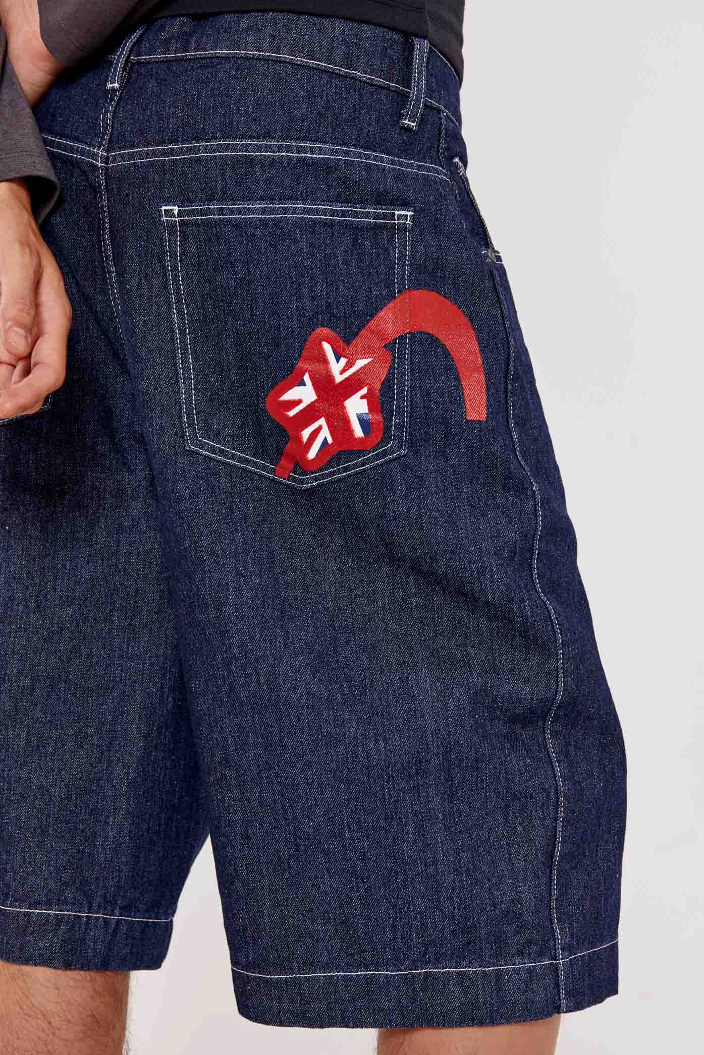 Male wearing indigo blue denim jorts in a jumbo fit with JL embroidered logo and Union Jack start branding on back pocket. 