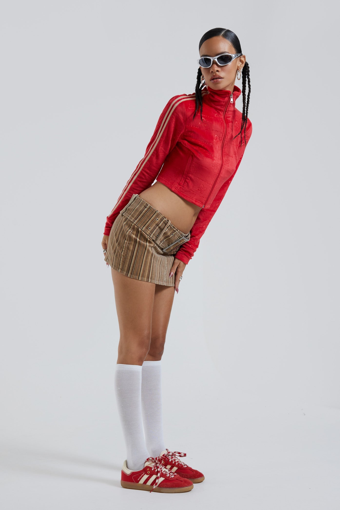 Female wearing red logo monogram embossed high neck long sleeve velour track top. Styled with brown stripe cord mini skirt. 