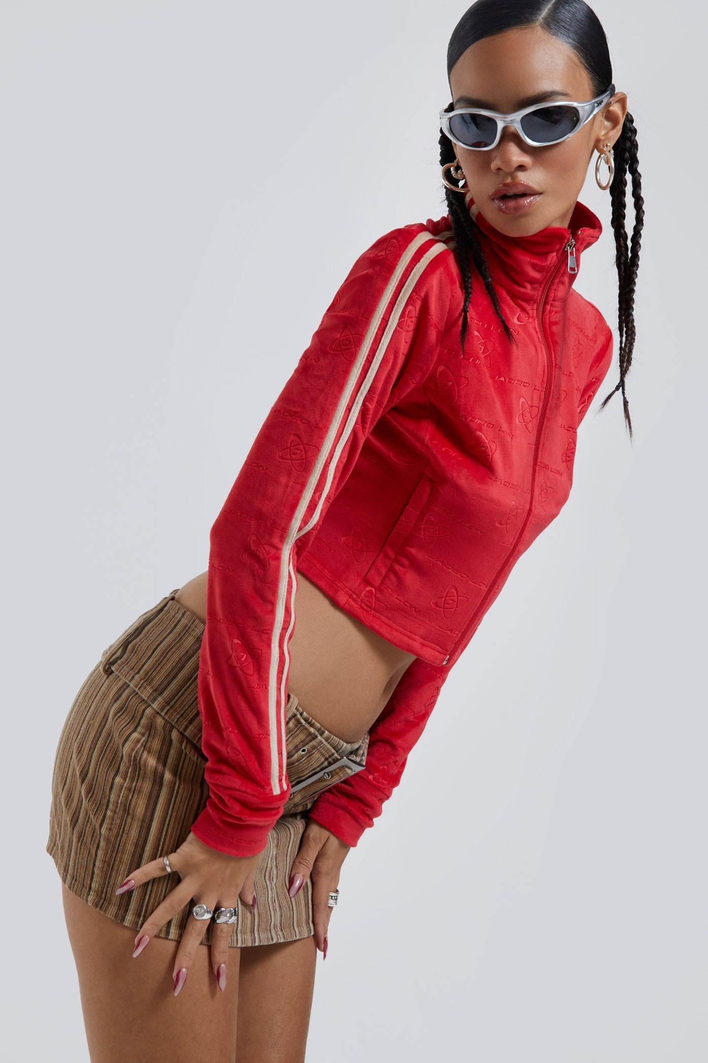 Female wearing red logo monogram embossed high neck long sleeve velour track top. Styled with brown stripe cord mini skirt. 