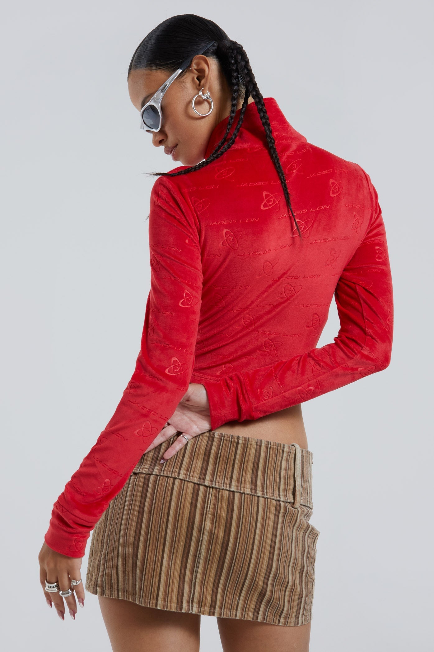 Female wearing red logo monogram embossed high neck long sleeve velour track top. Styled with brown stripe cord mini skirt. 