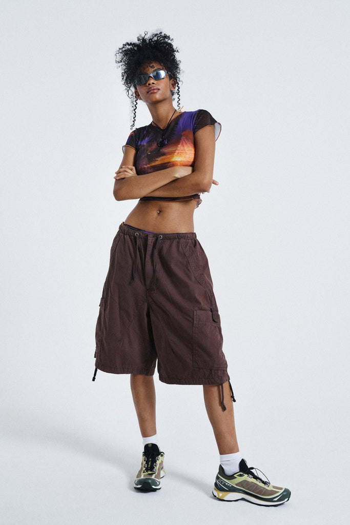 Female wearing Sunset Print Vintage Mesh Baby Tee. Styled with brown oversized parachute shorts.