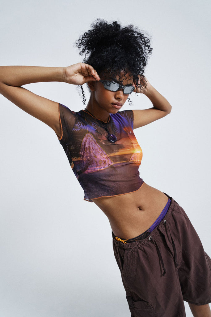 Female wearing Sunset Print Vintage Mesh Baby Tee. Styled with brown oversized parachute shorts.