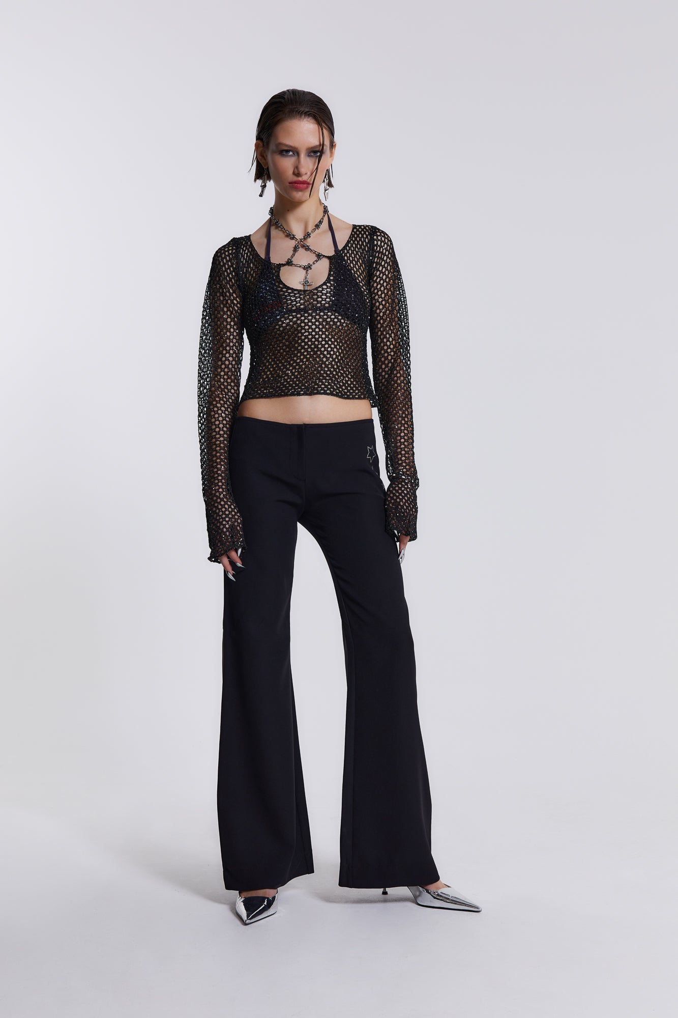 Female model wearing These black low waist trousers are in a wide leg fit and feature a silver diamante star detail.  Paired with a black crochet long sleeve top. 