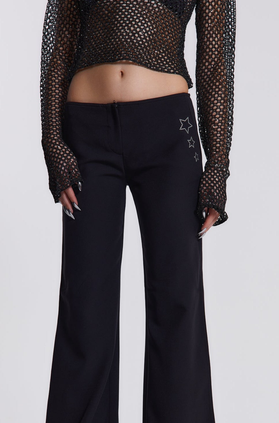 Female model wearing These black low waist trousers are in a wide leg fit and feature a silver diamante star detail.  Paired with a black crochet long sleeve top. 